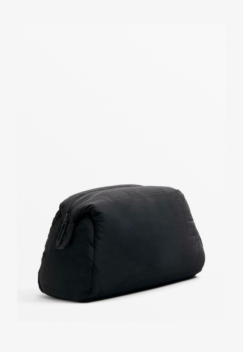 Borsetta nera rettangolare con zip e bordi arrotondati, realizzata in tessuto morbido, appoggiata su una superficie bianca.