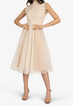 Ärmelloses beiges Spitzenkleid mit hohem Kragen und Tüllrock, getragen mit transparenten hochhackigen Knöchelriemchen-Schuhen.