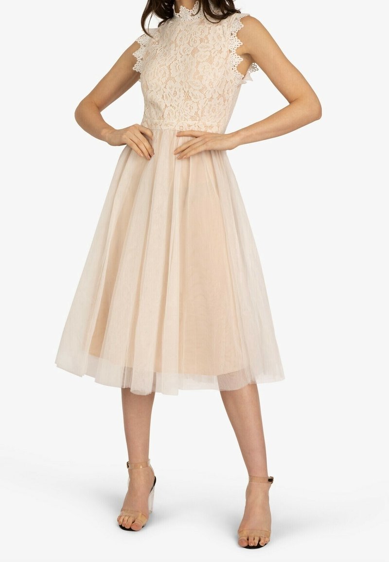 Robe sans manches en dentelle beige avec col haut et jupe en tulle, portée avec des chaussures transparentes à talons hauts et bride à la cheville.