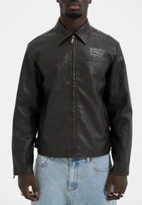Brown leather jacket with a collared neck, stitch detailing, and a silver zipper. Features white embroidered logo on the left chest.