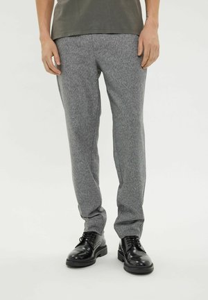 Man draagt grijze gestructureerde broek en zwarte gepolijste leren veterschoenen, staand tegen een effen lichte achtergrond.