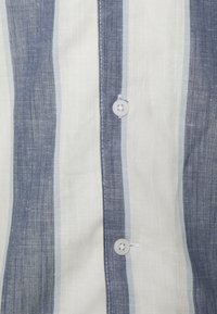 Camicia di lino con righe verticali blu e bianche, con bottoni sul davanti e tessuto leggero e strutturato, adatta per il clima caldo.