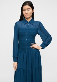 Tealfärgad klänning med långa ärmar, knäppning framtill, rynkad överdel och kjol i våder. Tyget verkar lätt och luftigt med en subtil glans.