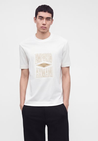 Νέος άνδρας που φοράει λευκό t-shirt Emporio Armani και μαύρο παντελόνι, στέκεται με τα χέρια στις τσέπες σε μονόχρωμο φόντο.