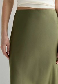 Falda de satén verde oliva con una textura suave y una silueta recta. Presentada con las manos posicionadas en la cintura.