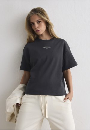 Pitkähiuksinen vaalea nainen, jolla on yllään tummanharmaa t-paita ja vaalean maitokermanväriset verkkarit, pitää käsissään maitokerman väristä takkia ja seisoo valkoista seinää vasten.