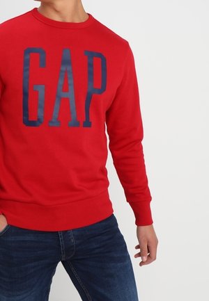 Person iført en rød langærmet sweatshirt med store marineblå bogstaver "GAP" og mørkeblå jeans mod en ensfarvet hvid baggrund.