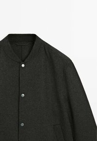 Dark grey wool jacket featuring a rounded collar, snap buttons, and side pockets. The fabric has a soft, textured appearance with a subtle sheen.