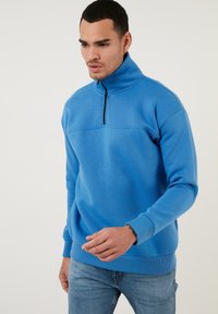 Pullover in pile blu con colletto alto e metà zip, caratterizzato da maniche e orlo a coste. Texture liscia e design minimalista.