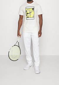 Camiseta blanca de algodón con un diseño gráfico amarillo y negro, combinada con pantalones deportivos blancos y zapatillas blancas, sosteniendo una raqueta de tenis.