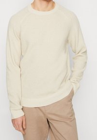 Beige texturerad stickad tröja med långa ärmar, rund halsringning och ribbade muddar och fåll. Matchad med ljusbeiga byxor.