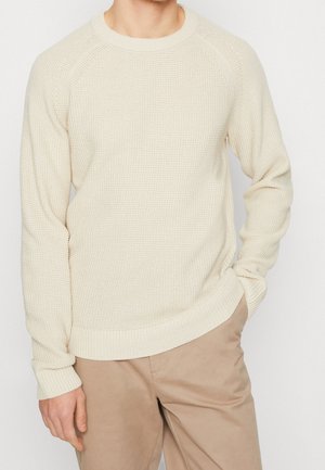 Pull en tricot texturé beige avec des manches longues, un col rond, et des poignets et un ourlet côtelés. Associé à un pantalon beige clair.