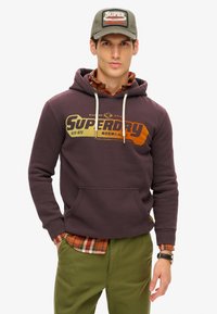 Ciemnofioletowa bluza z kapturem z logo "Superdry" w kolorze pomarańczowym i żółtym. Nałożona na koszulę w kratę, zielone spodnie i szary czapkę z naszywką.