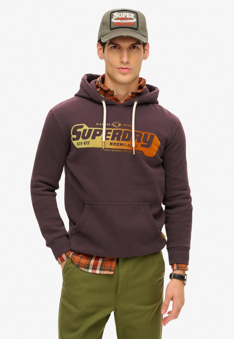 Ciemnofioletowa bluza z kapturem z logo "Superdry" w kolorze pomarańczowym i żółtym. Nałożona na koszulę w kratę, zielone spodnie i szary czapkę z naszywką.