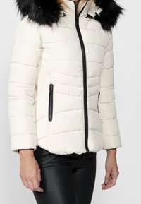 Giacca puffer trapuntata color crema con cappuccio in pelliccia nera, dotata di una zip frontale e tasche laterali diagonali. Pantaloni neri simili alla pelle.