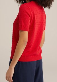 Röd stickad tröja med korta ärmar, strukturerade prickar överallt och ribbad fåll. Bärs över mörkblå byxor, visad från en sidovinkel.