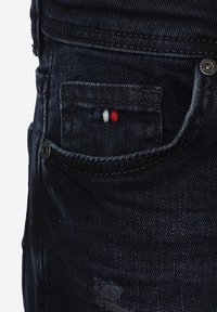 Jeans de mezclilla azul oscuro con un pequeño bolsillo frontal decorado con costuras en rojo, blanco y azul. Tejido texturizado con un detalle desgastado.
