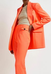 Klar orange kostymjacka med skräddarsydd passform, breda slag och framfickor, tillsammans med matchande högmidjade byxor och en ribbad beige topp.