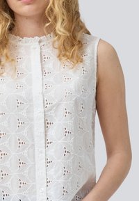 White sleeveless blouse with intricate cut-out floral pattern, button-down front, and ruffled collar detail. Lightweight fabric texture.