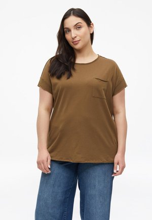 Femme aux longs cheveux foncés portant une chemise marron à manches courtes avec poche et un jean bleu, debout devant un fond blanc uni.