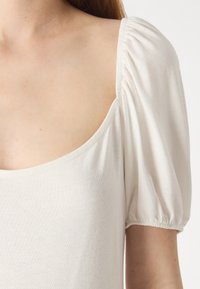 Haut blanc à encolure carrée, avec des manches bouffantes et un tissu doux et lisse. Le design présente de légères fronces aux bords des manches.