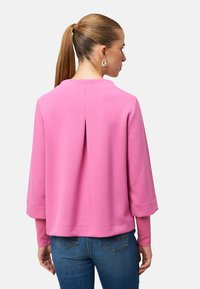 Pull-over rose avec un col rond et un pli à l'arrière. Manches longues avec poignets resserrés. Tissu doux et texturé. Associé à un jean bleu.