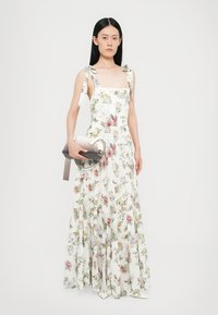 Floral maxi dress with square neckline, thick straps tied at shoulders, in white fabric with pink and green botanical print. Worn with a grey handbag.