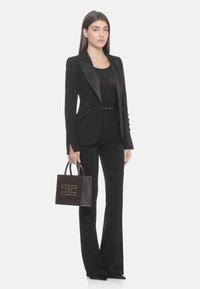 Abito nero su misura con rever in raso, top nero aderente e pantaloni svasati. Tiene una piccola borsa marrone con un logo dorato.