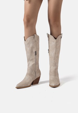 Gambe con stivali cowboy in camoscio beige a metà polpaccio con cuciture decorative e tacchi impilati in legno su sfondo bianco.