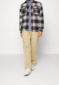 Giacca camicia in flanella scozzese nera, bianca e viola, con due tasche sul petto, indossata sopra una camicia azzurro chiaro con bottoni, abbinata a pantaloni beige e scarpe da ginnastica bianche.