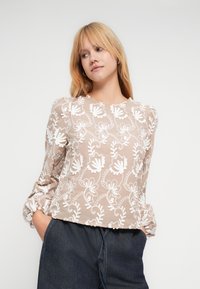 Moteris su blondinėmis plaukais, dėvinti smėlio spalvos palaidinę su baltais gėlių siuvinėjimais ir tamsiai mėlynas džinsines kelnes, rankos kišenėse, žiūrinti į šoną.