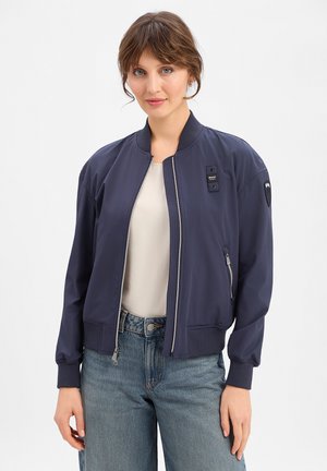 Frau mit hellbraunen Haaren trägt eine marineblaue Bomberjacke über einem weißen Oberteil und blauen Jeans und steht vor einem schlichten hellen Hintergrund.