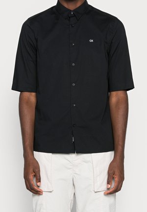 Homme portant une chemise noire à manches courtes boutonnée avec le logo "CK" sur la poitrine et un short cargo beige clair.