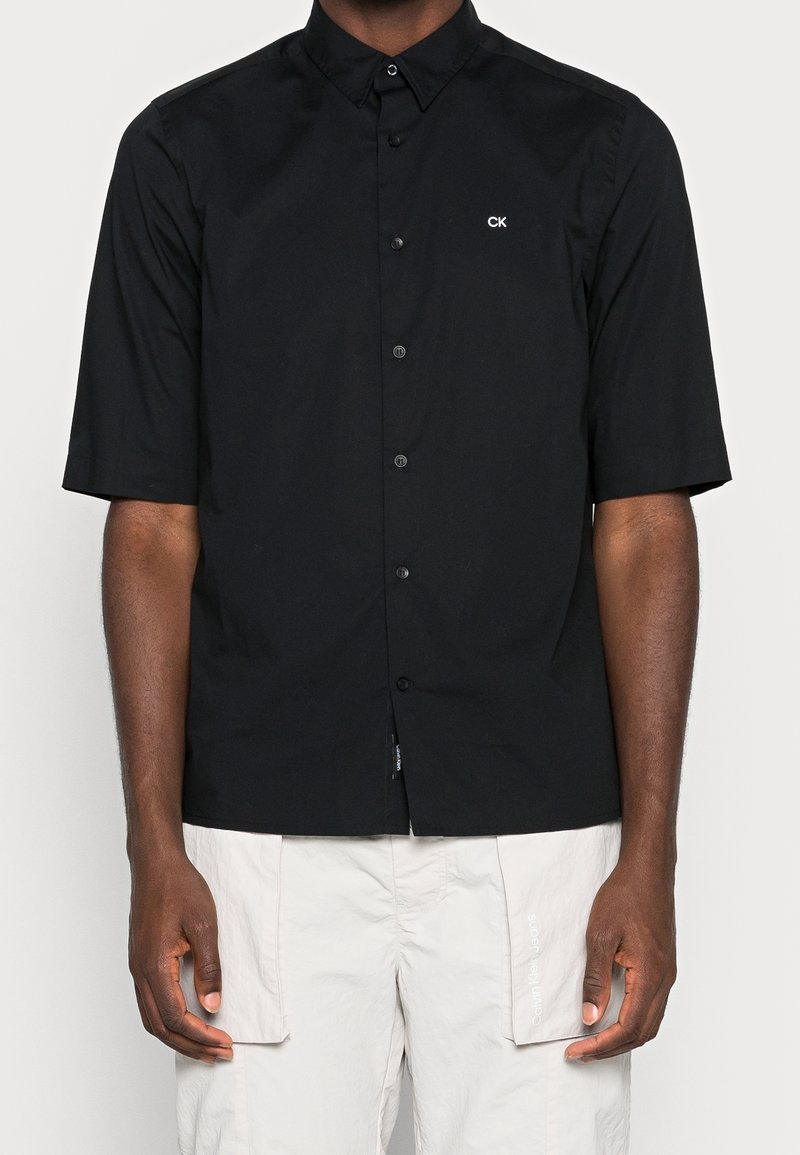 Homme portant une chemise noire à manches courtes boutonnée avec le logo "CK" sur la poitrine et un short cargo beige clair.