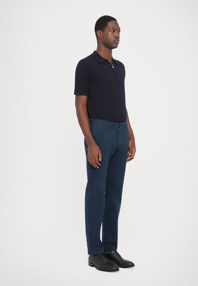 Navy polo shirt with short sleeves, made of lightweight fabric. Paired with dark navy trousers and black lace-up shoes, standing on a plain background.