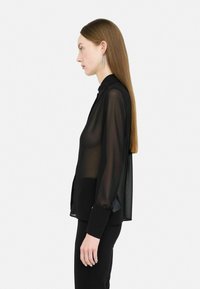 Blusa nera trasparente con una vestibilità rilassata, maniche lunghe e colletto con bottoni. Tessuto morbido e drappeggiato, abbinata a pantaloni neri.