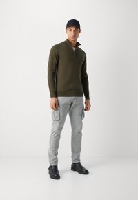 Veste zippée verte olive, pantalon cargo gris avec poches latérales, casquette noire et bottines noires, se tenant contre un fond blanc uni.