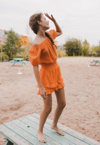 Oranje off-the-shoulder top met korte fladdermouwen en bijpassende gelaagde shorts, beide gemaakt van gestructureerde stof, gedragen op blote voeten op een zandachtige ondergrond.