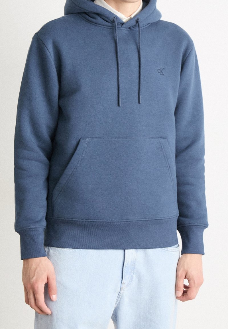 Personne portant un sweat à capuche bleu avec cordons et poche avant, affichant un petit logo « "CK" », assorti à un jean bleu clair.