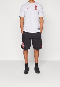 Muž oblečený v šedé košili s krátkým rukávem a černých šortkách s logy Bayern Mnichov a Adidas, stojící s rukama v kapsách.