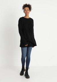 Schwarzer gerippter Oversize-Pullover mit langen Ärmeln, kombiniert mit schlanken blauen Jeans und schwarzen Schnürstiefeln. Glatte Textur, lockere Passform.