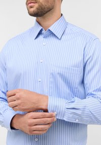 Camicia azzurra chiara con sottili strisce verticali bianche, colletto button-down e maniche lunghe. Presenta una tasca sul lato sinistro e polsini con bottoni.