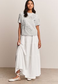 Grijze short-sleeve top met bloemenbroderie, gecombineerd met een witte gelaagde maxi rok. Licht gestructureerde stof en platte schoenen met enkelband.