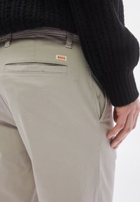 Lysegrå bukser lavet af bomuldsblanding, med et elegant design, sidelommer og et lille rødt og hvidt "BOSS"-mærke bagpå.