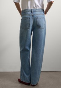 Persona in piedi con le mani sui fianchi, che indossa jeans larghi blu chiaro e una camicia bianca, davanti a una parete bianco sporco.