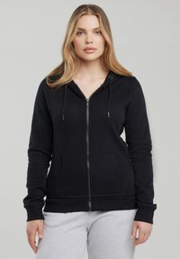 Kobieta o długich blond włosach, ubrana w czarną bluzę z kapturem na suwak i jasnoszare spodnie dresowe na jednolitym szarym tle.