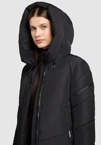 Frau in einer schwarzen gepolsterten Winterjacke mit großer Kapuze, zip und knopf geschlossen, vor einem schlichten hellgrauen Hintergrund.