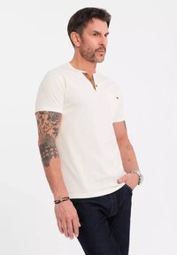 Cremefarvet kortærmet T-shirt med tre knapper og brystlomme. Har en tekstureret stof med brune detaljer.