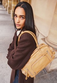 Hnědý koženkový satchel s několika zipovými přihrádkami, s hladkým povrchem a pevným držadlem. Nosený přes hnědý kabát.