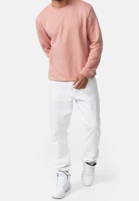 Jasnoróżowa bluza z okrągłym dekoltem, długimi rękawami i subtelnym logo; zestawiona z białymi dżinsami i białymi sneakersami z czarnymi akcentami.